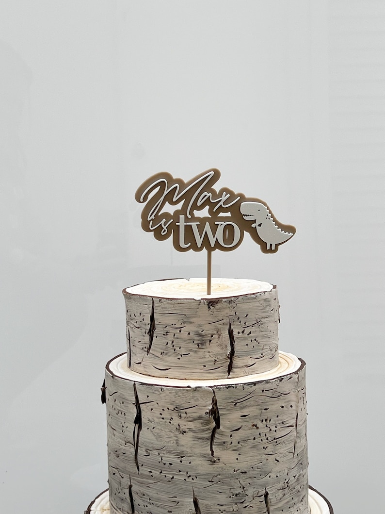 May include: A two-tiered cake decorated with a brown and white birch bark design. A brown and white cake topper with the text "Max is two" and a small white dinosaur is on top of the cake.
