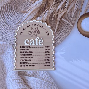 Puede incluir: Un letrero de madera con forma de arco caprichoso y un men&uacute; de cafeter&iacute;a. El letrero est&aacute; pintado de marr&oacute;n y tiene una inscripci&oacute;n blanca que dice "Bienvenido al caf&eacute; de Charlie". El men&uacute; enumera varios art&iacute;culos con precios, incluyendo babycino, cupcake, pan de hadas, batido de leche, rodajas de fruta, helado, galleta y huevos con tostadas.