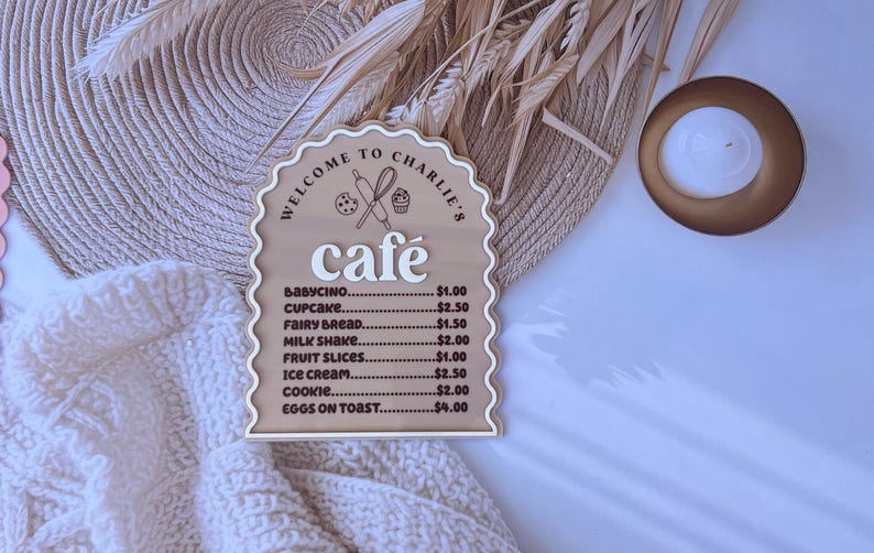 Puede incluir: Un letrero de madera con forma de arco caprichoso y un men&uacute; de cafeter&iacute;a. El letrero est&aacute; pintado de marr&oacute;n y tiene una inscripci&oacute;n blanca que dice "Bienvenido al caf&eacute; de Charlie". El men&uacute; enumera varios art&iacute;culos con precios, incluyendo babycino, cupcake, pan de hadas, batido de leche, rodajas de fruta, helado, galleta y huevos con tostadas.