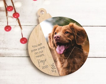 May include: Wooden Christmas ornament with a photo of a brown dog. The ornament has a diagonal split, with the left side featuring text that reads "You left paw prints on our hearts Kenzie 2020" and a leaf design.