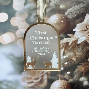 May include: A gold-colored Christmas ornament with the text "First Christmas Married" and the names "Mr & Mrs Samalra 2024". The ornament features deer and tree silhouettes. It hangs from a gold ribbon against a blurred Christmas tree background.