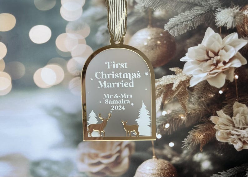 May include: A gold-colored Christmas ornament with the text "First Christmas Married" and the names "Mr & Mrs Samalra 2024". The ornament features deer and tree silhouettes. It hangs from a gold ribbon against a blurred Christmas tree background.