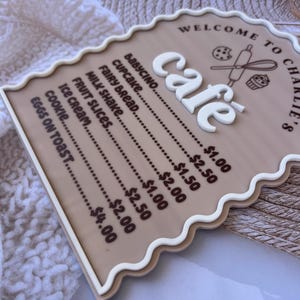 Puede incluir: Un letrero marr&oacute;n y blanco con un borde festoneado que dice "Bienvenido a Charlie's Cafe". El letrero enumera los art&iacute;culos del men&uacute; de la cafeter&iacute;a y sus precios, incluyendo babycino, cupcake, pan de hadas, batido de leche, rodajas de fruta, helado, galleta y huevos con tostadas.