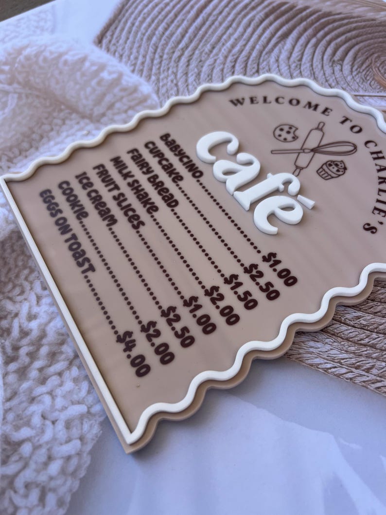 Puede incluir: Un letrero marr&oacute;n y blanco con un borde festoneado que dice "Bienvenido a Charlie's Cafe". El letrero enumera los art&iacute;culos del men&uacute; de la cafeter&iacute;a y sus precios, incluyendo babycino, cupcake, pan de hadas, batido de leche, rodajas de fruta, helado, galleta y huevos con tostadas.
