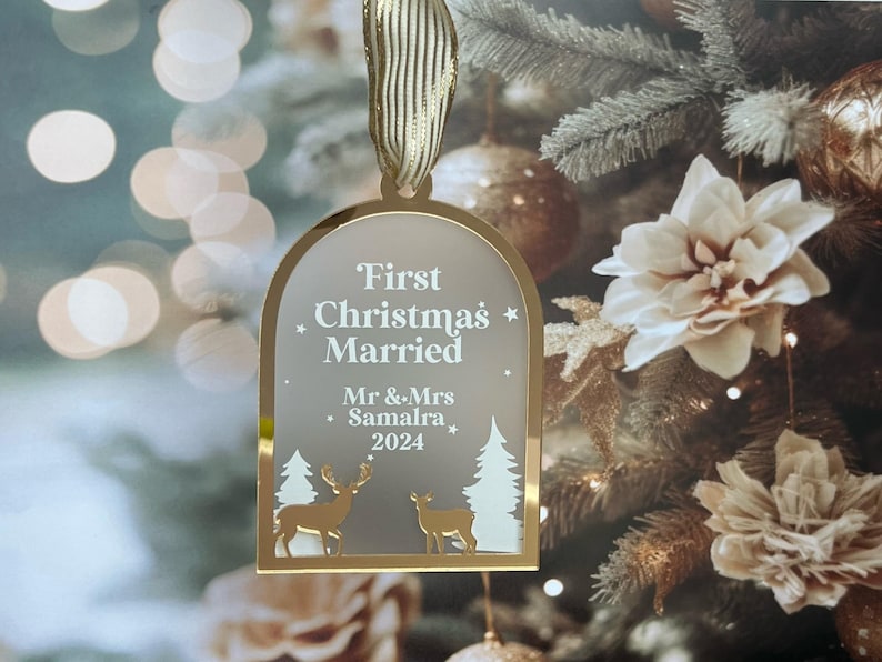 May include: A gold-colored Christmas ornament with the text "First Christmas Married" and the names "Mr & Mrs Samalra 2024". The ornament features deer and tree silhouettes. It hangs from a gold ribbon.