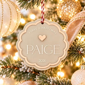 May include: A scalloped, beige circular Christmas ornament hanging from a red and white string. The ornament features the text "FIRST CHRISTMAS", a small heart icon, the name "PAIGE", and the year "2026" in raised, light-colored lettering.