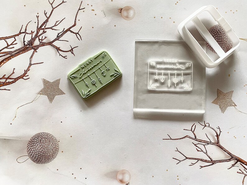 May include: Two Christmas themed rubber stamps, one with the text "Merry Christmas" and a garland of ornaments, the other with a garland of ornaments. The stamps are on a white surface with a few decorative branches and gold glitter.