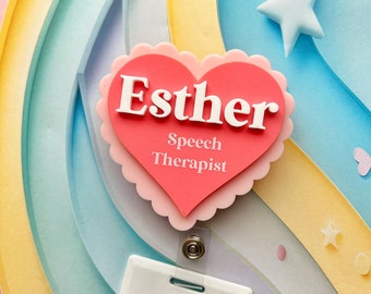 May include: A pink heart-shaped name tag with the name "Esther" and the text "Speech Therapist" in white letters. The name tag is attached to a white badge holder with a silver clip.