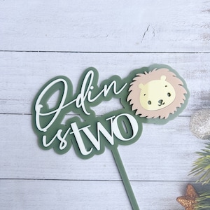 May include: A green and white cake topper with the words "Odin is two" in a script font. A cartoon lion face is on the right. The topper is made of layered acrylic and is designed for a birthday cake.