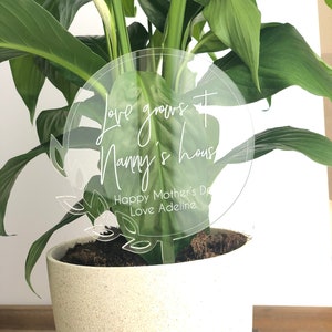May include: A white acrylic plant stake with the text "Love grows at Nanny's house. Happy Mother's Day Love Adeline" in a circle with a leafy design.