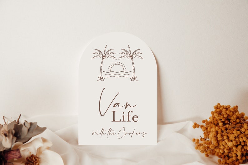May include: White arch-shaped sign with a brown line drawing of two palm trees, a sun setting over the ocean, and the text "Van Life with the Crokers".