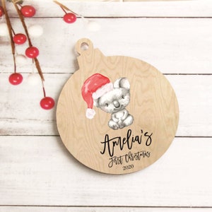 Puede incluir: Adorno navide&ntilde;o de madera con forma de bola, con un koala con gorro de Pap&aacute; Noel. El adorno marr&oacute;n claro tiene el texto "Amelia's First Christmas 2020" en escritura negra.
