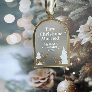 May include: A gold-colored Christmas ornament with the text "First Christmas Married" and the names "Mr & Mrs Samaira 2024". The ornament features deer and tree cutouts, and is hanging from a gold ribbon. The background is a blurred Christmas tree.