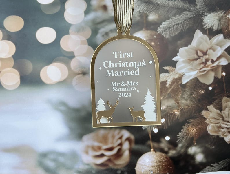 May include: A gold-colored Christmas ornament with the text "First Christmas Married" and the names "Mr & Mrs Samaira 2024". The ornament features deer and tree cutouts, and is hanging from a gold ribbon. The background is a blurred Christmas tree.