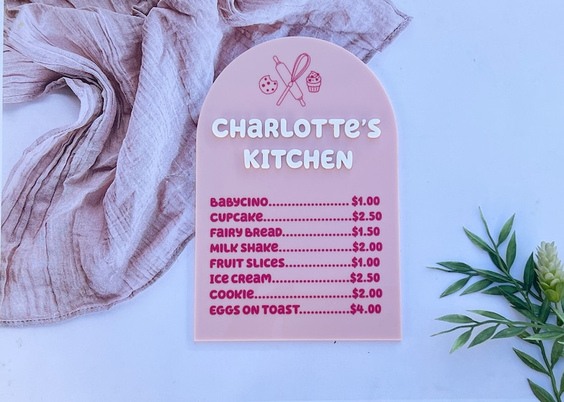 Pode incluir: Uma placa rosa com um arco branco e o texto "CHARLOTTE'S KITCHEN" em branco. A placa lista um menu de alimentos para crian&ccedil;as com pre&ccedil;os, incluindo babycino, cupcake, fairy bread, milk shake, fatias de fruta, sorvete, biscoito e ovos com torradas.