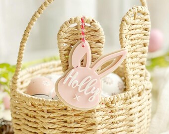 May include: A pink bunny-shaped Easter basket tag with the name "Holly" written in white. The tag is hanging from a woven wicker basket with bunny ears.
