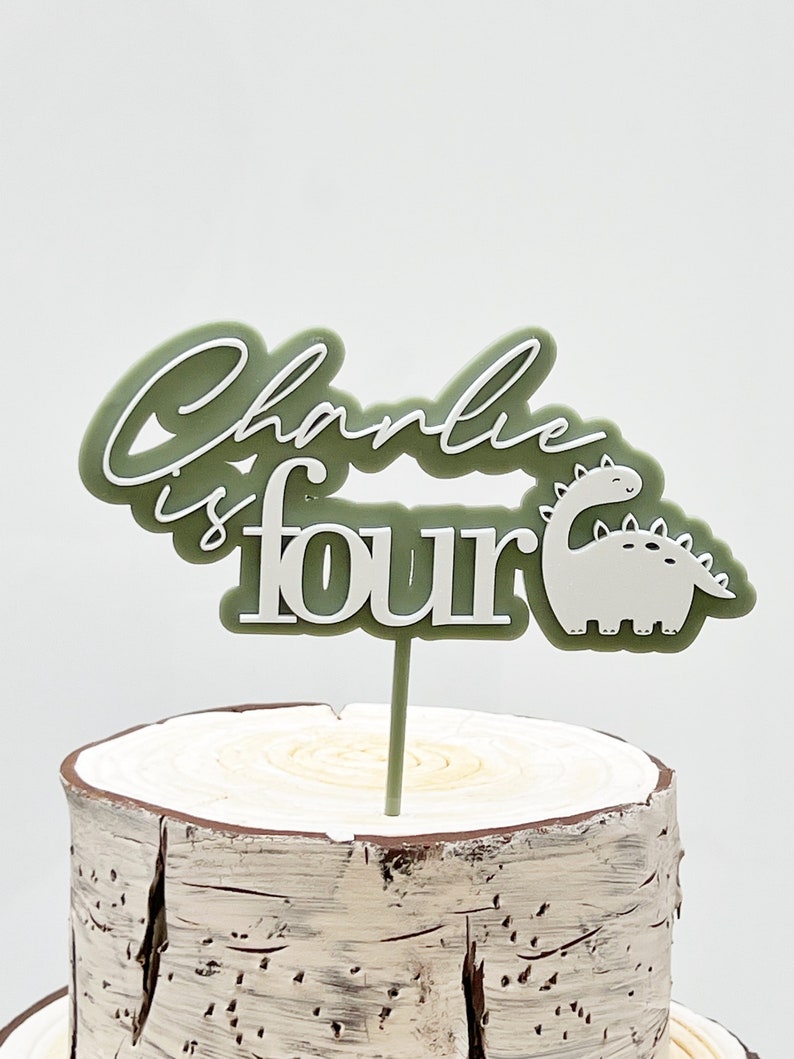 May include: Green and white cake topper with the text "Charlie is four" and a white dinosaur illustration.