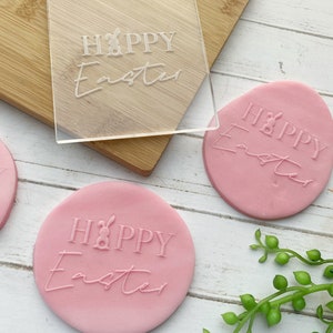 May include: A clear acrylic stamp with the text "HAPPY Easter" and a bunny rabbit graphic. The stamp is used to create impressions on pink clay circles with the same text.
