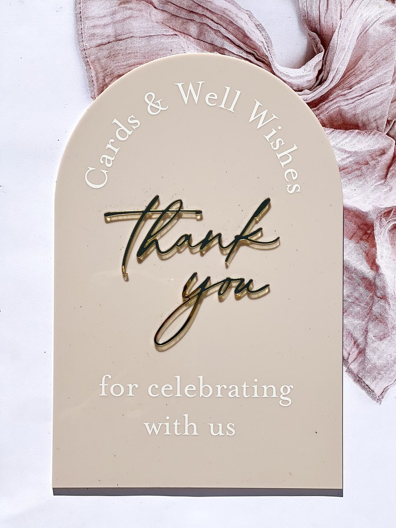 K&ouml;nnte beinhalten: Ein beigefarbenes Schild in Bogenform mit dem Text "Cards & Well Wishes" oben und "Thank you for celebrating with us" unten. Die Worte "Thank you" sind in goldener Schrift geschrieben.