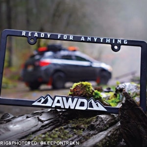 May include: Black license plate frame with the text "READY FOR ANYTHING" and a white mountain range graphic with the letters "AWD" inside. The frame is sitting on a mossy log with a black car in the background.