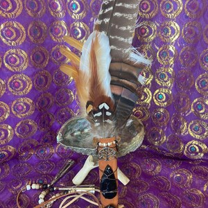 May include: A fan made of layered brown, white, and tan feathers attached to a tan leather handle. The handle features a silver tree charm, two black stones wrapped in dark wire, and a dangling cord with white, brown, and clear beads.