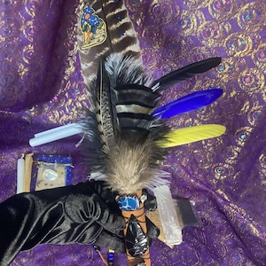 May include: A ceremonial fan featuring a large brown and white striped feather with a wolf and moon patch, layered with black, blue, and yellow feathers. The handle is wrapped in tan leather with a blue stone, black obsidian shards, and black cord.