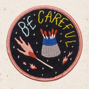 May include: A black and white embroidered patch with a pink hand holding a matchstick. The matchstick is lit and the flame is red. The patch has the text "BE CAREFUL" in yellow and blue lettering.