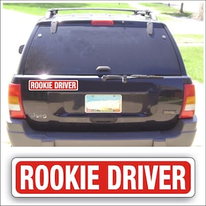 May include: A red and white rectangular sign with the text "ROOKIE DRIVER" in white letters. The sign is attached to the back of a black Jeep.