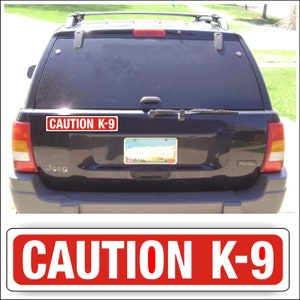 May include: A red and white rectangular sticker with the text "CAUTION K-9" is affixed to the rear of a dark blue Jeep. The sticker is above the license plate. The image also shows a close-up of the sticker.