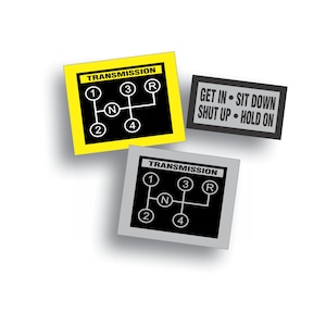 May include: Three yellow and gray stickers with black text and graphics. The stickers show a car transmission gear shift pattern with the letters 'R', 'N', '1', '2', '3', and '4'. The text on the stickers reads 'TRANSMISSION' and 'GET IN - SIT DOWN - SHUT UP - HOLD ON'.