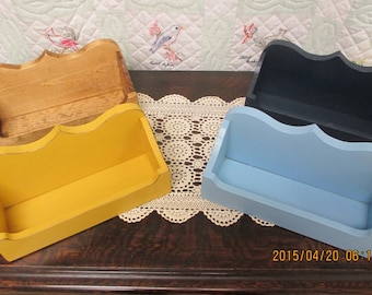 May include: Four wooden wall shelves in different colors: natural wood, yellow, light blue, and dark blue. The shelves have a decorative curved top edge and are arranged on a wooden surface.