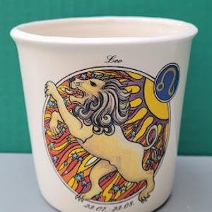 May include: White ceramic planter with a colorful illustration of a lion with the zodiac symbol for Leo and the text "Leo" and "32 oz.  A108".