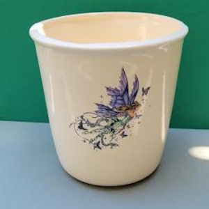 Puede incluir: Una maceta de cerámica blanca con un diseño de un hada morada con alas y mariposas.