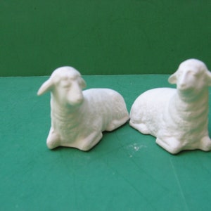 May include: Two white ceramic sheep figurines lying down on a green surface. The sheep have detailed features and are ready to be painted.