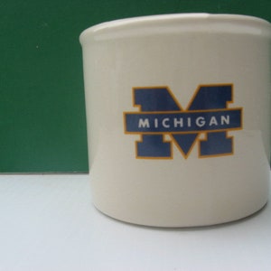 May include: White ceramic jar with a blue and gold Michigan logo. The logo is a large "M" with the word "Michigan" underneath.