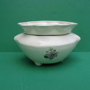 May include: A white ceramic two-piece serving bowl with a floral design. The bottom bowl has a rounded shape with three small feet. The top piece is a smaller, scalloped-edged dish. The floral design features purple flowers and green leaves.