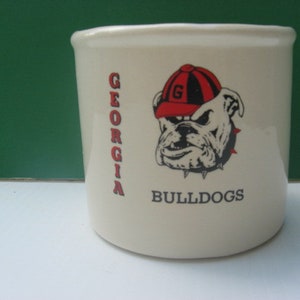 May include: Cream-colored ceramic mug featuring the text "GEORGIA" in red vertical lettering and the word "BULLDOGS" in black. The mug displays a bulldog mascot wearing a red and black cap.