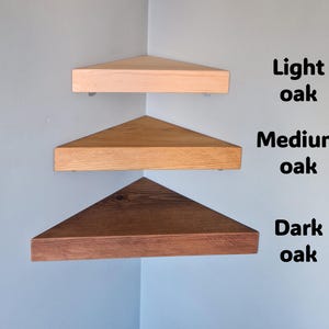 Op de afbeelding: Drie houten hoekplanken in lichte, medium en donkere eikentinten zijn aan een lichtblauwe muur bevestigd. De planken zijn driehoekig, met de tekst "Light oak", "Medium oak" en "Dark oak" naast elke plank.