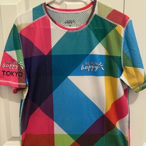 May include: A colorful running shirt with a geometric pattern in shades of blue, green, pink, yellow, and white. The shirt has the text "WE RUN HAPPY" on the front and "WE RUN HAPPY TOKYO" on the left sleeve.