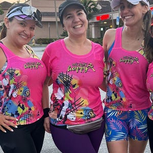Puede incluir: Tres mujeres con camisetas de running rosas con el texto "We Run Happy" en amarillo. Las camisetas tienen un diseño abstracto colorido. Dos mujeres llevan leggings negros y morados, y una mujer lleva pantalones cortos con estampado azul y blanco.