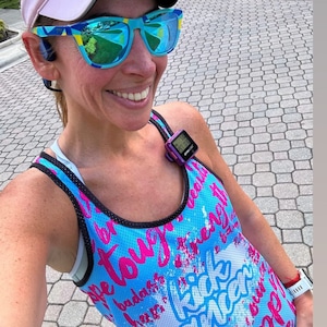 May include: A woman wearing a light blue visor, blue and yellow sunglasses, and a blue tank top with pink text. The tank top has the words "Kick Cancer" and other phrases. A purple fitness tracker is attached to the tank top.