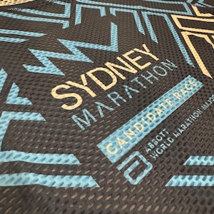 May include: A black running shirt with a blue and white geometric pattern. The shirt has the text "Sydney Marathon" and "CandidaTe Race" printed on it. The text "Abbott World Marathon Majors" is also printed on the shirt.