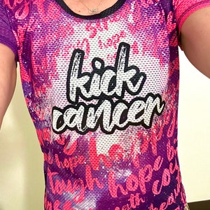 May include: A pink and purple athletic shirt with a mesh fabric design. The shirt has the words "kick cancer" written in white on the front. The shirt is being worn by a person.