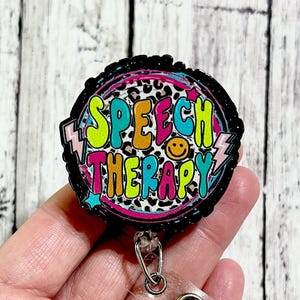 May include: A black and white leopard print circle badge with the text "SPEECH THERAPY" in colorful letters. The badge is attached to a clear retractable badge holder.
