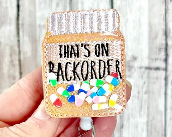 May include: A gold pill bottle with the text "That's on Backorder" and colourful pills inside. The bottle is attached to a clear plastic badge holder with a silver clip.