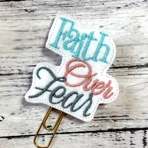 May include: White felt paper clip with the words "Faith Over Fear" embroidered in teal, pink, and gray thread.