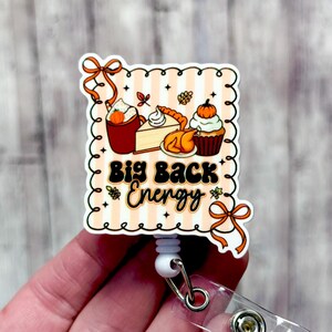 May include: A fall-themed badge reel with a scalloped edge and the text "Big Back Energy." The design includes illustrations of a pumpkin spice latte, pie, turkey, and a cupcake. The background has a peach and white striped pattern.