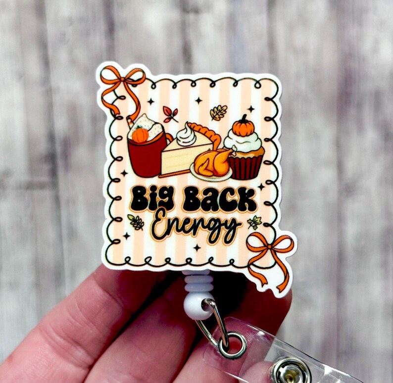May include: A fall-themed badge reel with a scalloped edge and the text "Big Back Energy." The design includes illustrations of a pumpkin spice latte, pie, turkey, and a cupcake. The background has a peach and white striped pattern.