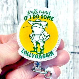 May include: A white and green glitter badge holder with a yellow circle and a cartoon frog wearing a hat. The text on the badge holder reads "Y'all mind if I do some lollygaggin'"