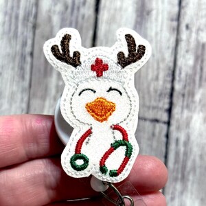 May include: A white and brown embroidered badge reel featuring a smiling cartoon character dressed as a nurse with reindeer antlers. The character has an orange beak, a red cross, and is holding a red and green stethoscope.